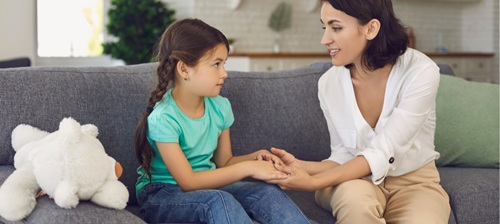 A woman and child sit on a sofa holding hands. The woman seems to be talking to the girl. To the left of the girl  on the sofa is a white stuffed toy.
