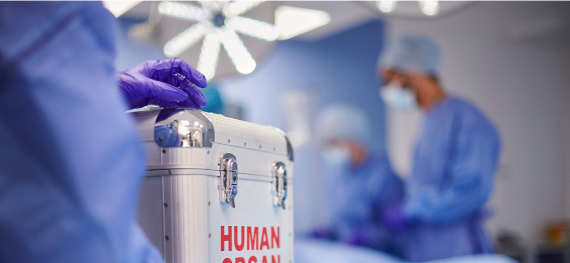 An operating theatre, blurred figures in the background. In the foreground a metal case with 'human organ' written in red on it.