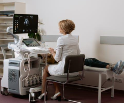 A radiologist sits on a chair with an ultrasound machine and screen in front of them. A person lies on a bed next to the radiologist, ready for their scan.