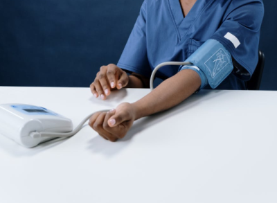 A picture showing a person having a blood pressure reading. They have a cuff wrapped around their upper arm. Their lower arm is resting on a table. A cable comes from the cuff to a device which displays their blood pressure.